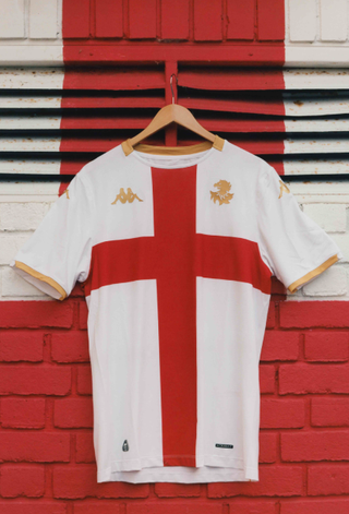 A white Genoa CFC away shirt featuring a large red St. George's cross, gold cuffs and collar trim, and a golden griffin crest, hanging on a wooden hanger.