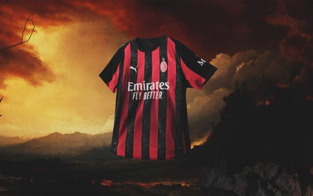 The AC Milan 2025/26 home shirt featuring classic red and black vertical stripes, presented against a dramatic fiery sky background.