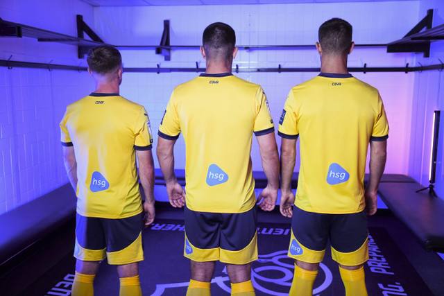 Three players seen from behind wearing the yellow Derby County 2024/25 third shirt in a locker room setting.