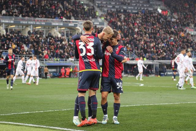 Bologna FC 1909 players celebrate on the pitch wearing their red and blue striped home shirt.