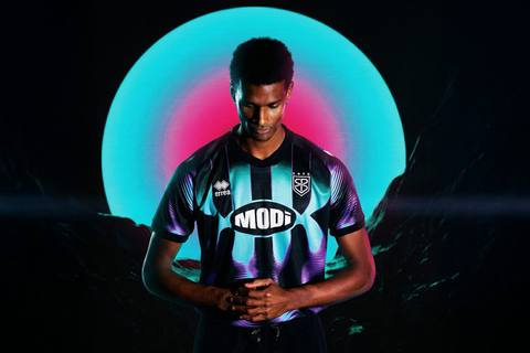 A model wears the new San Pio X away shirt by Erreà, featuring a black base with cyan and purple gradients.