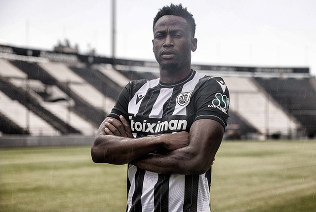 PAOK FC player posing in the 2024/25 home shirt with classic black and white vertical stripes on a stadium pitch.