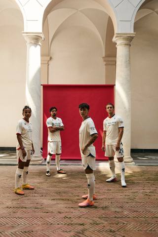 Models posing in a courtyard wearing the Genoa away kit, featuring a white base with red and blue details.