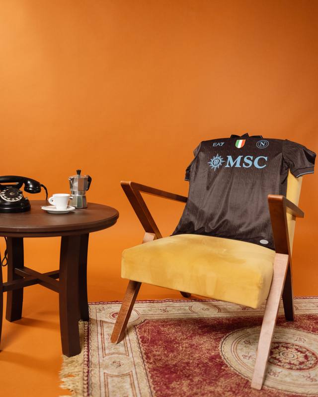 The SSC Napoli 2025/26 third shirt 'Café' is displayed draped over a yellow armchair in a retro coffee-themed setting.