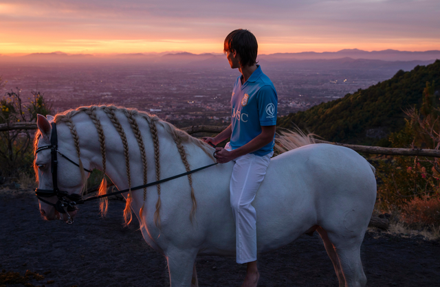 A campaign image showing a model on a white horse wearing the SSC Napoli home shirt at sunset.