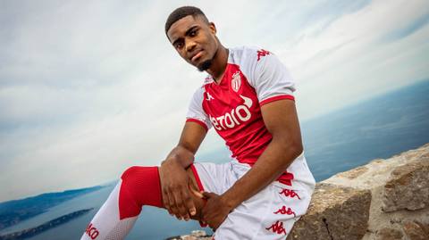 A player wearing the AS Monaco 2022/23 home kit by Kappa, sitting on a stone ledge overlooking the sea.