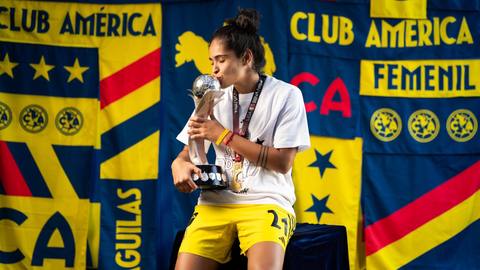 A Club América Femenil player kisses a championship trophy in front of club banners