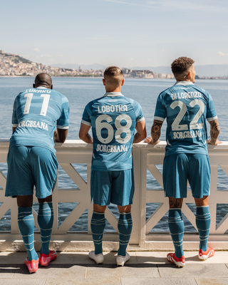 Three SSC Napoli players wearing the Partenope fourth kit are shown from behind, leaning against a white railing overlooking the sea.