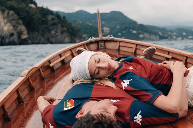 Two models wear the Genoa CFC 2024/25 home shirt while relaxing on a wooden boat.