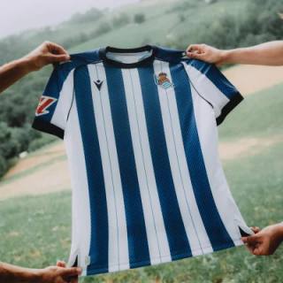 The Real Sociedad home shirt featuring classic blue and white vertical stripes, held up against a green hillside backdrop.