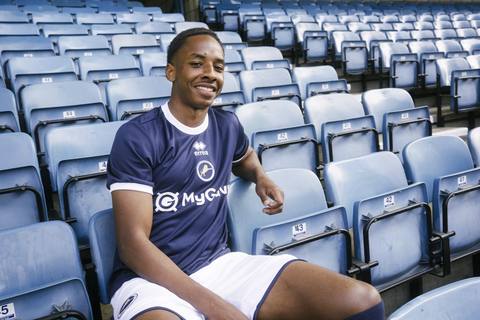 A model wears the Millwall FC 2025/26 home shirt while posing in the stadium stands.
