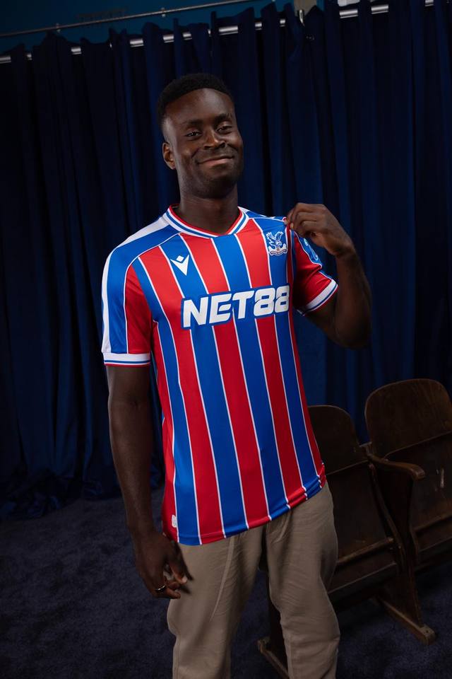 Marc Guéhi models the Crystal Palace FC 2025/26 home shirt, featuring red and blue stripes with white pinstripe borders.