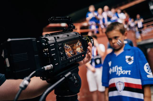 A behind-the-scenes photograph showing a video camera filming a young fan wearing the Sampdoria home shirt.