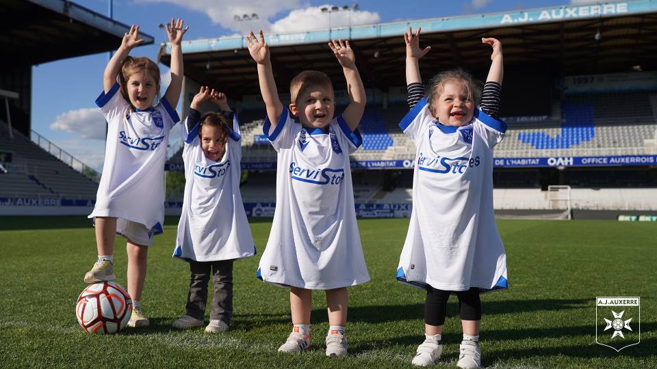 Four young children pose on the pitch at Stade de l'Abbé-Deschamps wearing AJ Auxerre's 2022/23 white and blue home shirt.