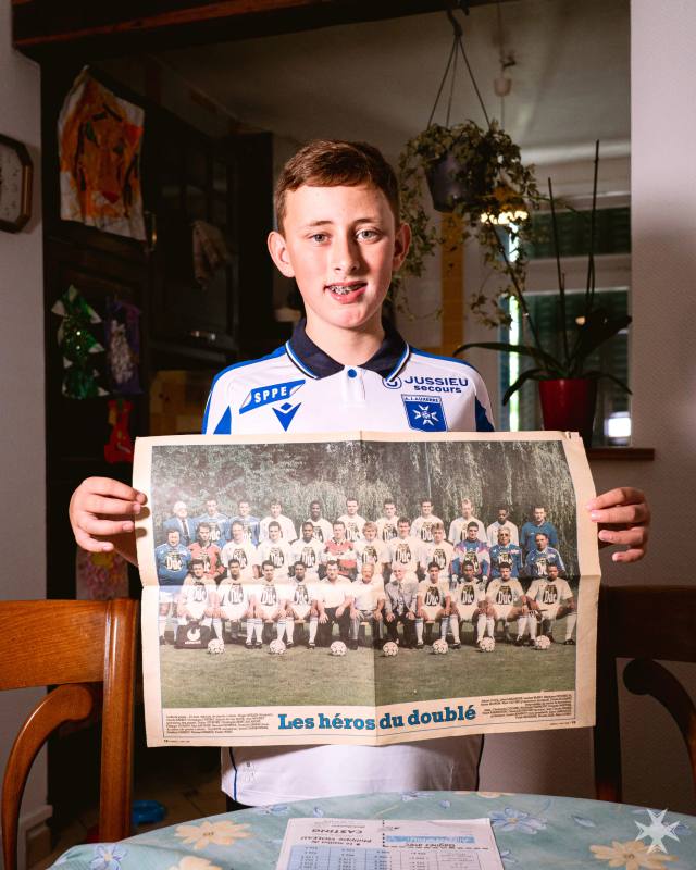 A young fan wearing the AJ Auxerre home shirt while holding a historic newspaper clipping showing the team.