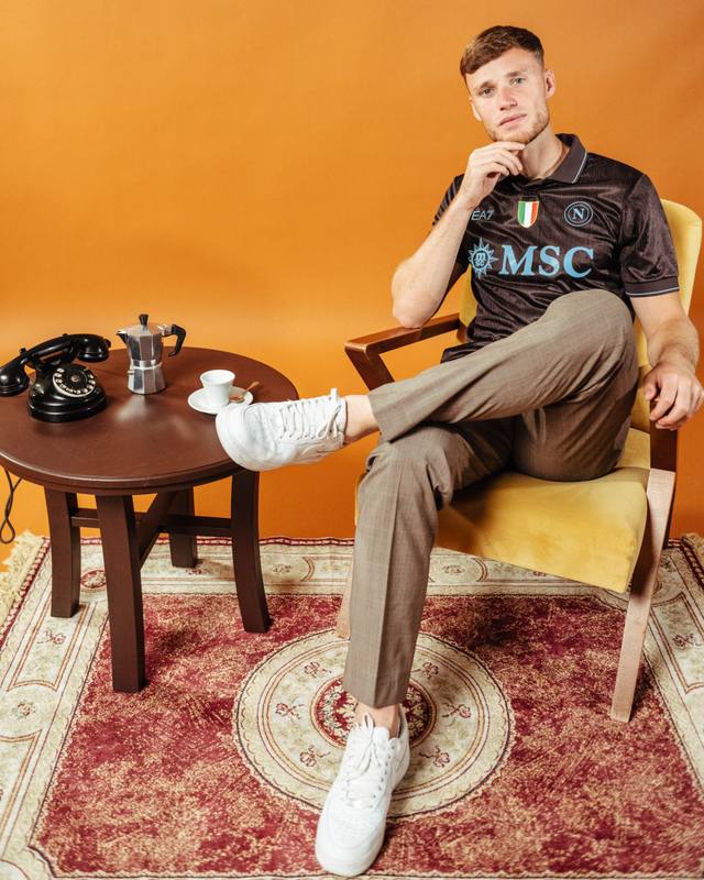 A player seated in a retro-styled living room set wears the dark brown SSC Napoli third shirt.
