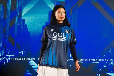 A model presents the Mansfield Town FC 2025/26 away shirt by Erreà, featuring a navy and blue graphic pattern.