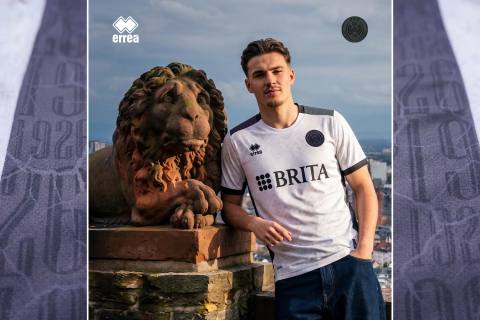 A model posing in the white SV Wehen Wiesbaden 2026/27 away shirt next to a stone lion statue.