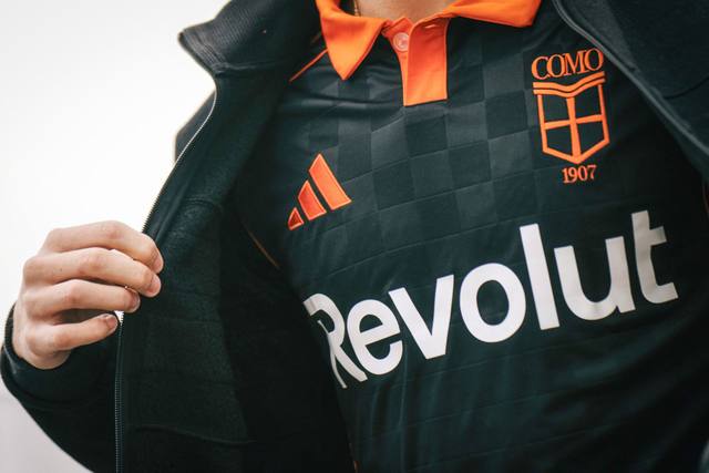 A close-up of the Como 1907 third shirt featuring a black checkered pattern and orange accents, shown under a black unzipped jacket.