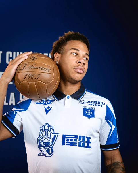 A player wearing the AJ Auxerre home shirt while holding a vintage leather football.