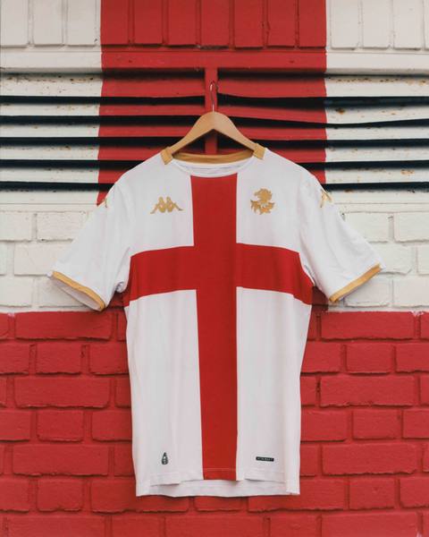 A white Genoa away shirt featuring a red St. George's cross, hanging on a wooden hanger against a red and white brick wall.